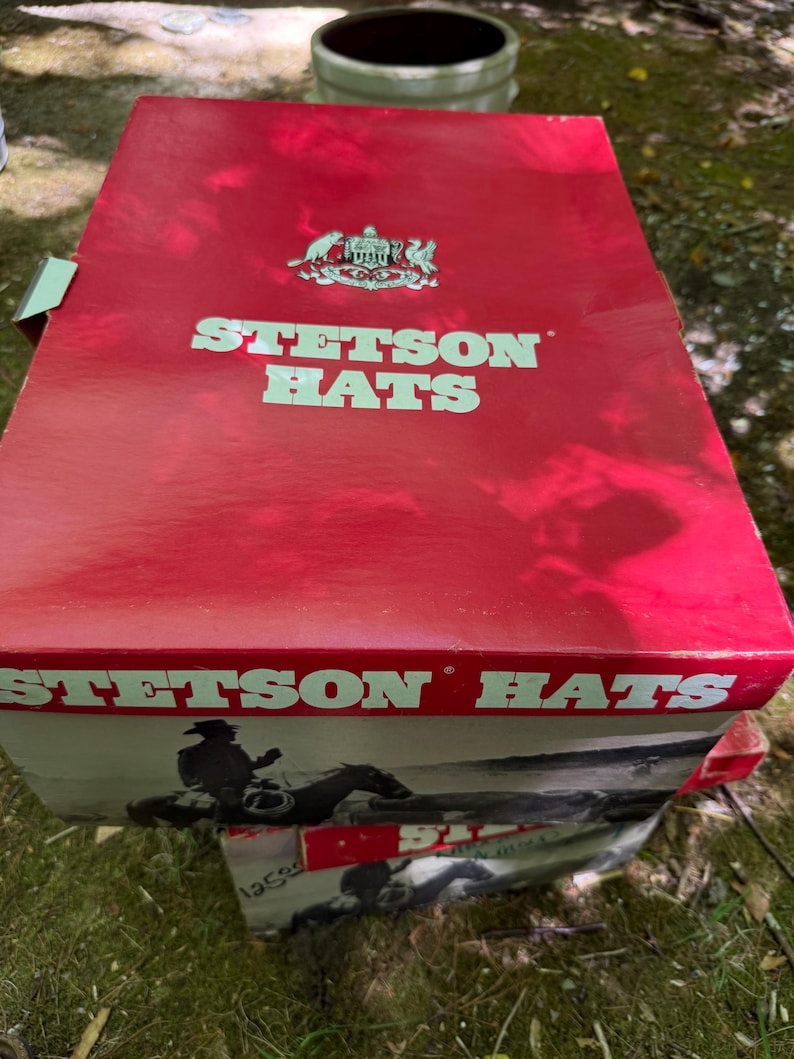 May include: A red Stetson Hats box with white lettering and a logo. The box features an image of a cowboy on a horse. The box is sitting on top of another box with a similar design.