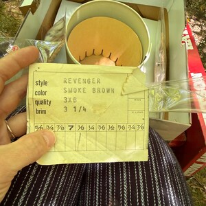 May include: An open box containing a hat and a label. The label reads "REVENGER SMOKE BROWN 3XB" with a brim size of 3 1/4 inches. The hat is inside a round container. The box is brown and white.