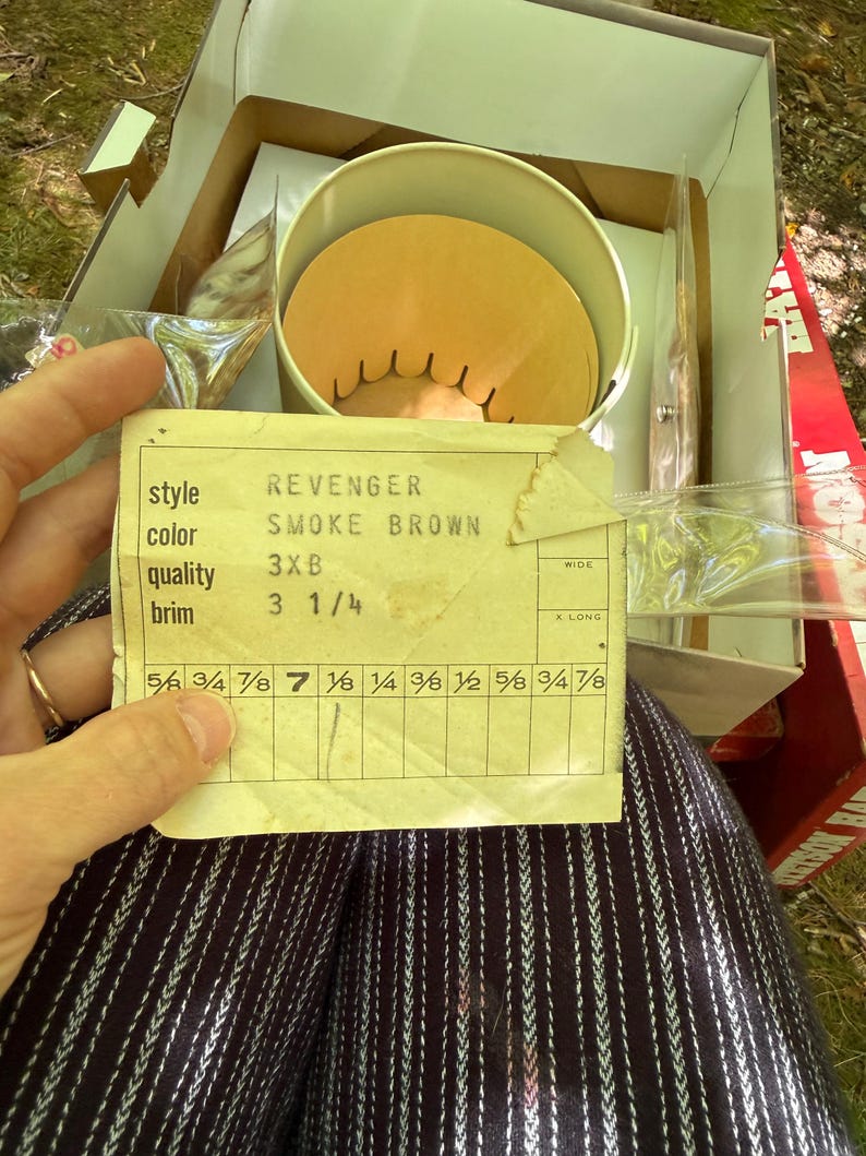 May include: An open box containing a hat and a label. The label reads "REVENGER SMOKE BROWN 3XB" with a brim size of 3 1/4 inches. The hat is inside a round container. The box is brown and white.