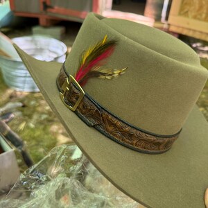 May include: A khaki-colored cowboy hat with a decorative leather band featuring a floral pattern and a gold-colored buckle. The hat is adorned with a cluster of colorful feathers, including red, yellow, and brown.