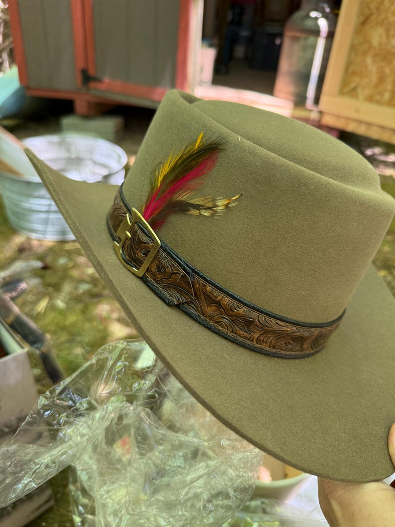 May include: A khaki-colored cowboy hat with a decorative leather band featuring a floral pattern and a gold-colored buckle. The hat is adorned with a cluster of colorful feathers, including red, yellow, and brown.