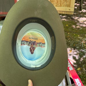 May include: A Stetson cowboy hat in a muted olive green color. The hat's interior features a colorful image and the Stetson logo. The brim is wide, and the hat appears to be made of felt or a similar material. The hat is being held by a person.