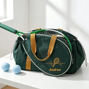 Peut inclure: Un sac de tennis vert foncé avec des accents dorés et une raquette de tennis à l'intérieur. Le sac porte un motif de raquette de tennis et le nom "Andrea". Deux balles de tennis bleu clair sont devant le sac.