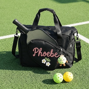 May include: A black sports bag with a pickleball paddle tucked into the front pocket. The bag features the name Phoebe embroidered in pink, a white flower appliqué, and a small green creature keychain. Two pickleballs, one lime green and one yellow, sit in front of the bag.