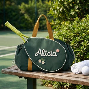 Op de afbeelding: Een donkergroene tennistas met mosterdgele handvatten en een witte bies. De tas is voorzien van de naam Alicia in witte schrijfletters, versierd met kleine witte en roze bloemapplicaties. Een limoengroen tennisracket zit in de tas gestoken.