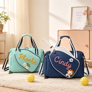May include: Two pickleball bags, one mint green and one navy blue, each featuring a front pocket shaped like a pickleball paddle. The mint bag has the name Phoebe embroidered in yellow, and the navy bag has the name Cindy in pink.