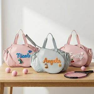 May include: Three sports bags in light pink and light blue, each featuring a name in raised, colourful lettering. The bags have side pockets and carry handles. A tennis racket is tucked into the centre bag, and three pink tennis balls are nearby.