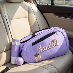 Peut inclure: Un sac de sport violet clair portant le nom Nandini en lettres texturées en relief et de petites fleurs brodées. Le sac est doté d'une fermeture éclair noire et de poignées noires. À côté se trouvent une gourde violette et une balle de tennis.