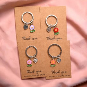 Op de afbeelding: Vier sleutelhangers met bloembedels in roze, rood, oranje en paars, elk bevestigd aan een zilveren sleutelring. Elke sleutelhanger heeft een hartvormige bedel met de tekst "Thank You". De sleutelhangers worden gepresenteerd op bruine kaarten met de tekst "Thank You" en "For Helping Me Grow".