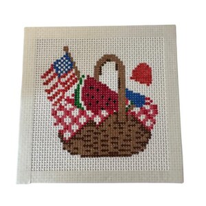 Può includere: Un pezzo quadrato di tela plastica bianca con un disegno a punto croce che raffigura un cestino da picnic marrone riempito con un panno a quadri rossi e bianchi, una fetta di anguria, una piccola bandiera americana e un oggetto rosso.