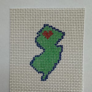 May include: A square piece of white needlepoint canvas featuring a cross-stitched outline of the state of New Jersey in dark blue thread. The interior of the state shape is filled with light green thread and includes a small red heart.