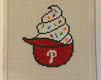 May include: A square needlepoint canvas featuring a cross-stitched design of a soft-serve ice cream cone inside a red baseball cap. The cap displays a white letter P logo on the front, and the ice cream is topped with small colorful sprinkles.