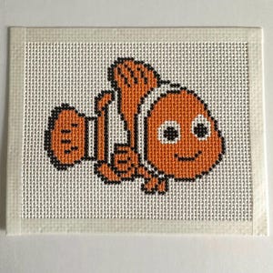 May include: A needlepoint canvas featuring a cross-stitched clownfish design. The fish is orange with white vertical stripes and black outlines, rendered in a pixelated style on a white grid mesh fabric with taped edges for embroidery projects.
