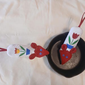 May include: Two white oven mitts with red mouse heads and red hanging loops. Each mitt has a floral design with a red tulip, green leaves, and a blue vase. The mitts are next to a black cast iron skillet.