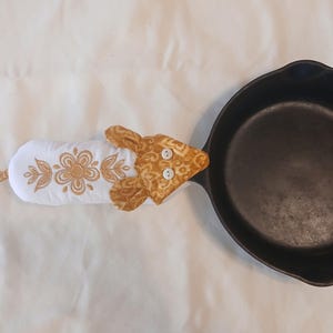 May include: A black cast iron skillet with a white and gold floral-embroidered oven mitt. The oven mitt has a tan patterned head and two white buttons. The mitt is attached to the skillet.