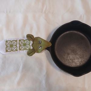 May include: A black cast iron skillet with a white and green decorative pot holder. The pot holder has a white base with green floral embroidery and a green frog-like head. The skillet is round and has a single handle.