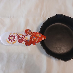 May include: A black cast iron skillet with a white and red floral patterned oven mitt. The mitt features a bird and flower design with red, yellow, and orange accents. The skillet is empty.