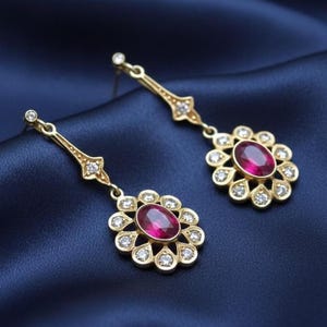 May include: A pair of gold-toned drop earrings with a floral design. Each earring features a central oval ruby surrounded by small, clear gemstones. The earrings hang from a decorative gold bar with additional small gemstones. The background is a dark blue fabric.