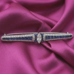 May include: A long, silver brooch with a central marquise-cut diamond. The brooch features a design of square blue gemstones and smaller white gemstones. The brooch is set against a pink satin background.