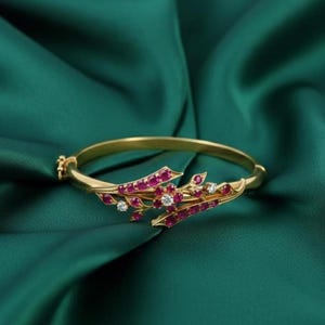 Könnte beinhalten: Ein goldfarbenes Armband mit floralem Design. Das Armband hat rote Edelsteine und kleine weiße Edelsteine. Das Armband ist vor einem dunkelgrünen Stoffhintergrund platziert.