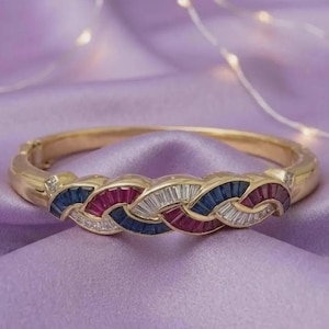 Könnte beinhalten: Goldfarbenes Armband mit geflochtenem Design. Das Armband hat abwechselnde Abschnitte aus roten, weißen und blauen Edelsteinen. Das Armband befindet sich auf einem lila Hintergrund.