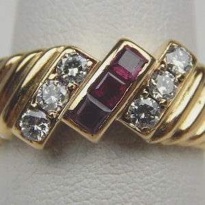 Può includere: Un anello d'oro con tre diamanti trasparenti su ciascun lato di un'incastonatura rettangolare con tre pietre preziose quadrate rosso scuro. L'anello ha una fascia a coste.
