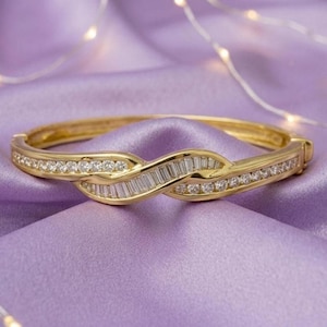 Puede incluir: Un brazalete rígido articulado en tono dorado con diseño cruzado. La superficie está incrustada con una hilera de pequeñas piedras redondas transparentes y una sección central de piedras rectangulares en talla baguette.