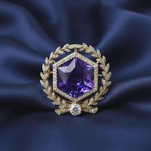 May include: A gold brooch featuring a large, hexagonal, deep purple gemstone at the centre, surrounded by a frame of small, clear, sparkling stones. The hexagonal setting is encircled by a detailed gold laurel wreath design with small clear stones.