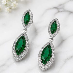 May include: A pair of drop earrings featuring two marquise-cut emerald green stones in each piece, surrounded by a halo of small clear white stones set in a silver-toned metal frame with a hinged connection between the two stone sections.
