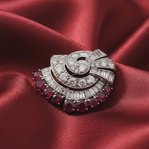 Op de afbeelding: Een zilverkleurige broche met een spiraalvormig ontwerp bezet met heldere ronde en baguette-geslepen stenen, geaccentueerd door een gebogen rij donkerrode ronde stenen langs de onderrand, wat een waaiervorm creëert met metalen details.