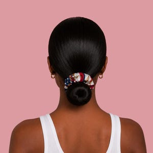 Op de afbeelding: Een haarelastiek met een rood, wit en blauw patroon van sterren en strepen, vastgemaakt rond een knot met donker haar. Het stoffen accessoire toont een patriottisch ontwerp met zichtbare witte sterren op een blauw veld en rode strepen.