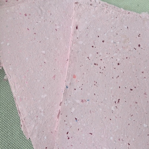 May include: Several sheets of light pink handmade paper with a rough, deckled edge. The paper has a textured surface embedded with small, scattered flecks of dark red, white, and blue material throughout, creating a speckled appearance.