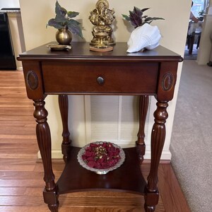 May include: A dark brown wooden side table with a drawer and a lower shelf. The table has ornate legs and decorative oval accents. A gold statue, a white seashell, and a small vase with plants are on top. A silver dish with red petals sits on the shelf.