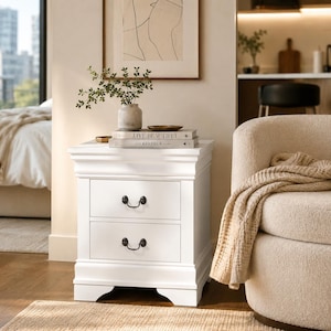 May include: A white nightstand with two drawers and black hardware. The nightstand has a decorative top and base. A vase with greenery, books, and a small dish sit on top. A beige armchair and a window are in the background.