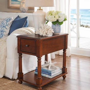 May include: A dark brown wooden side table with a single drawer and a lower shelf. The table has turned legs and a dark knob on the drawer. A white lamp, coral, and a vase of white flowers sit on top. The table is 30 inches long.