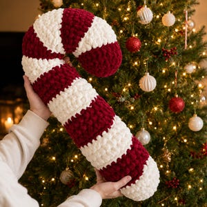 May include: A large, plush, crochet candy cane cushion featuring alternating thick stripes of deep burgundy red and creamy white yarn. The soft, textured fabric is crafted in a chunky knit style, forming a curved, hook-shaped decorative item.