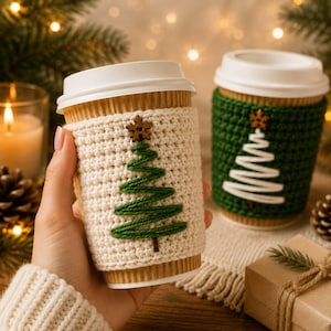 May include: Two disposable coffee cups with white lids, each featuring a textured crochet sleeve. One sleeve is cream-colored with a green yarn-stitched Christmas tree and a small wooden star button, while the other is dark green with a white tree.