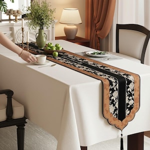 May include: A white tablecloth with a decorative table runner. The runner has a black center with floral patterns and a brown border. A cup and saucer are on the table, along with a bowl of green apples and a vase with greenery.
