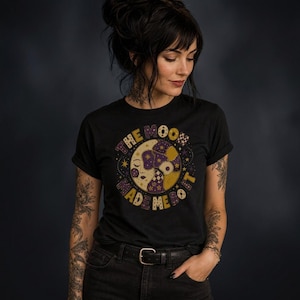 May include: A black short-sleeve t-shirt featuring a circular graphic of a crescent moon with a face. The design includes purple and gold patchwork patterns, small stars, and the text "THE MOON MADE ME DO IT" in a bold, arched font around the moon.