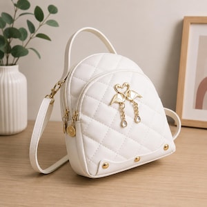 May include: A white quilted mini backpack featuring a top handle and adjustable shoulder straps. The front is adorned with a gold-toned metal flamingo emblem with dangling chain accents and teardrop charms, plus three gold-toned metal studs.