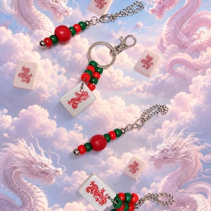 May include: A collection of four keychains featuring white mahjong tiles with red dragon engravings. Each keychain includes a silver metal ring or clasp, accented with alternating red and green spherical beads, and a small silver metal chain link.