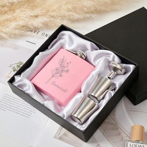 Könnte beinhalten: Ein rosafarbener, rechteckiger Metallflachmann mit einer schwarzen Strichzeichnung eines Blumenstraußes und dem Namen Samarah, präsentiert in einer schwarzen Geschenkbox mit weißem Satinfutter, inklusive zwei silbernen Metallbechern und einem Trichter.