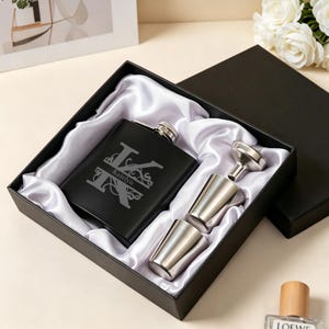 May include: A black matte stainless steel flask engraved with a large letter K and the name Katelyn, presented in a black gift box with white satin lining, including two silver-toned metal shot cups and a small silver-toned metal funnel.