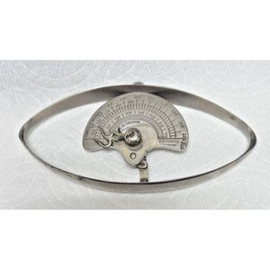 Könnte beinhalten: Vintage silberfarbenes Instrument mit ovalem Rahmen. In der Mitte befindet sich ein halbkreisförmiges Messgerät mit Gradmarkierungen und dem Text "DE PRESSION". Das Instrument hat ein Scharnierdesign und wird wahrscheinlich zur Messung verwendet.