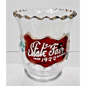 Op de afbeelding: Een heldere glazen votiefkaarsenhouder met een geschulpte rand en gouden rand. De houder heeft een rood label met de woorden "State Fair 1922" in witte letters en turquoise bloemaccenten.