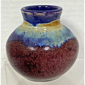 May include: A small, round ceramic vase with a wide, bulbous body and a narrow neck. The glaze features a deep, speckled maroon base that transitions into a vibrant light blue and tan drip pattern around the upper shoulder and the rim.