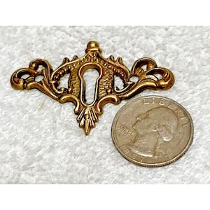 May include: Ornate brass keyhole cover with a decorative design. The cover is next to a quarter for size comparison. The keyhole cover has a detailed, vintage aesthetic.