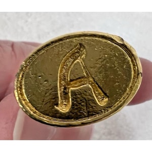 May include: A gold-colored, oval-shaped button or charm with a raised letter "A" in the center. The letter and the border around the oval are textured. The background is a light color.
