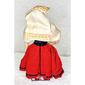 Peut inclure: Une poup&eacute;e vintage portant un foulard blanc brod&eacute; de rouge et de jaune, une jupe rouge avec une bordure noire et un corsage bleu. La poup&eacute;e tient un panier dans sa main droite.