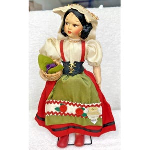 Peut inclure: Une poup&eacute;e vintage v&ecirc;tue d'une robe traditionnelle italienne avec un chemisier blanc, une jupe rouge et verte et un chapeau blanc. La poup&eacute;e tient un panier de fleurs violettes.